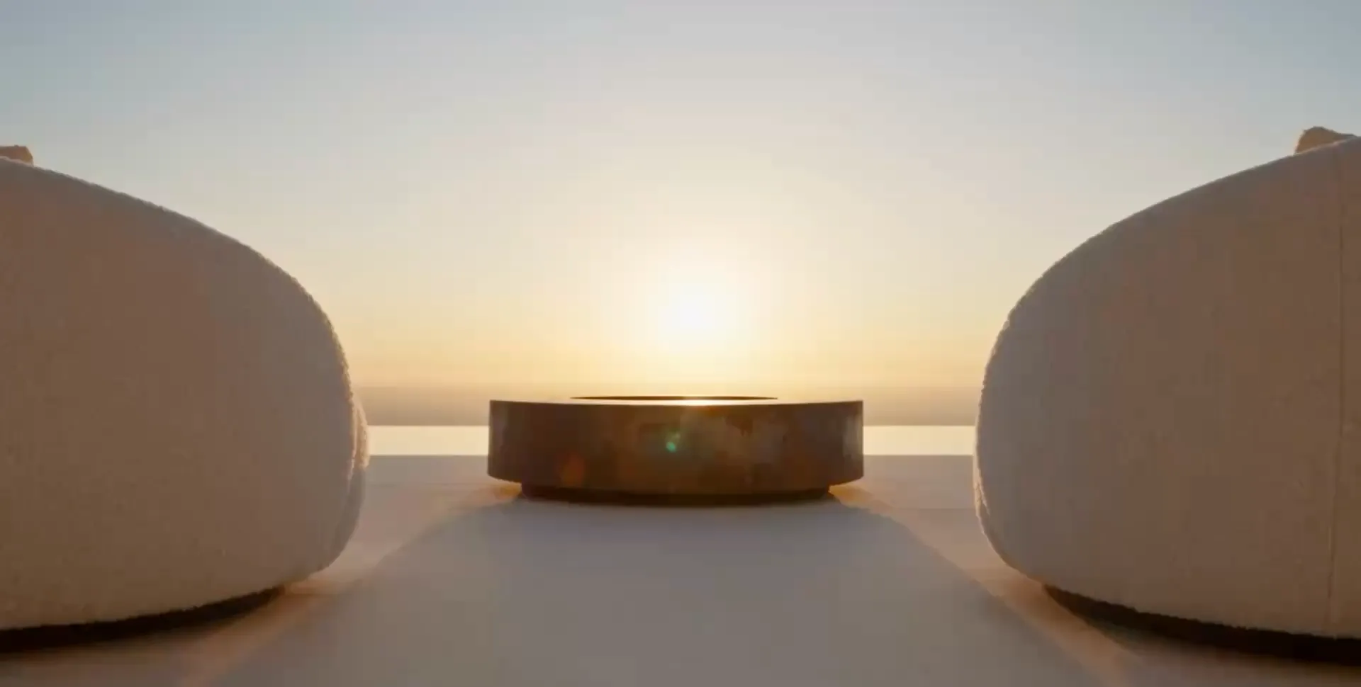 Hélios fire table in Corten steel facing the sun setting over the sea, between two blurred white armchairs